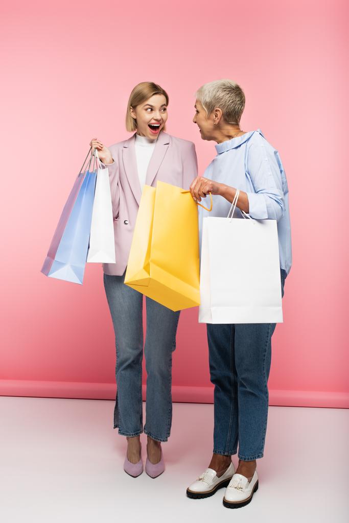 πλήρες μήκος των ενθουσιασμένοι νεαρή γυναίκα και ώριμη μητέρα σε τζιν κρατώντας τσάντες ψώνια σε ροζ - Φωτογραφία, εικόνα