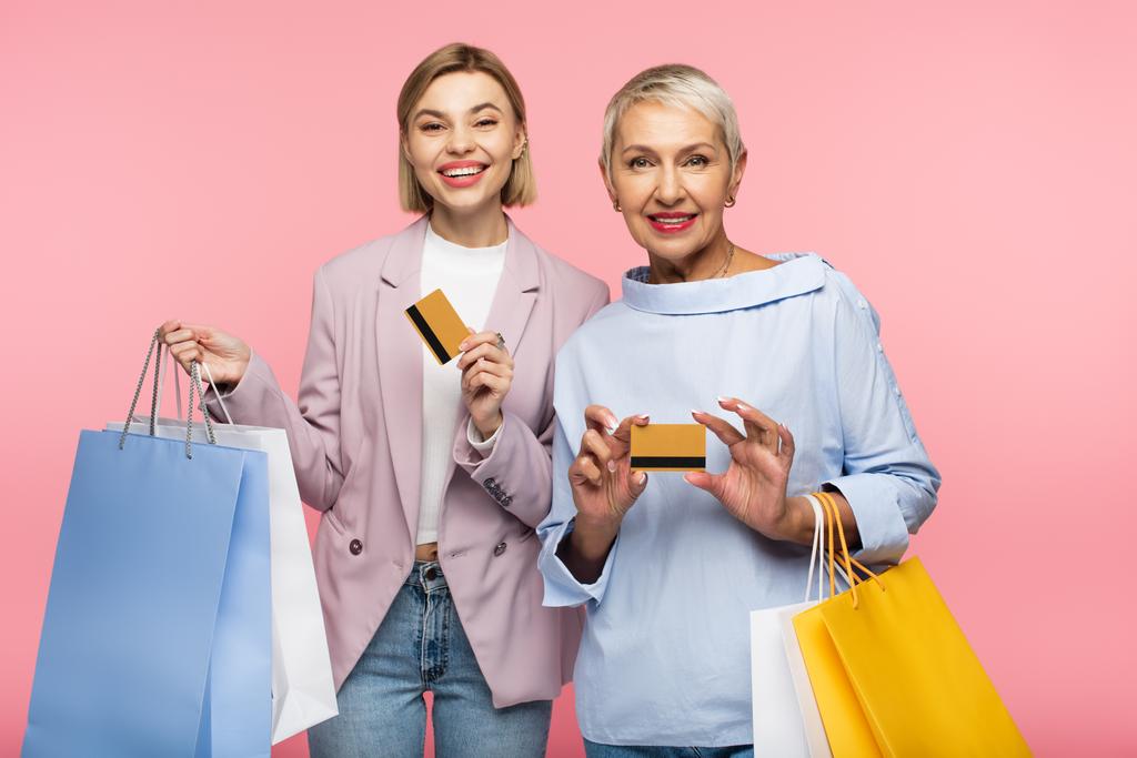 alegre joven y madura madre sosteniendo bolsas de compras y tarjetas de crédito aisladas en rosa  - Foto, imagen