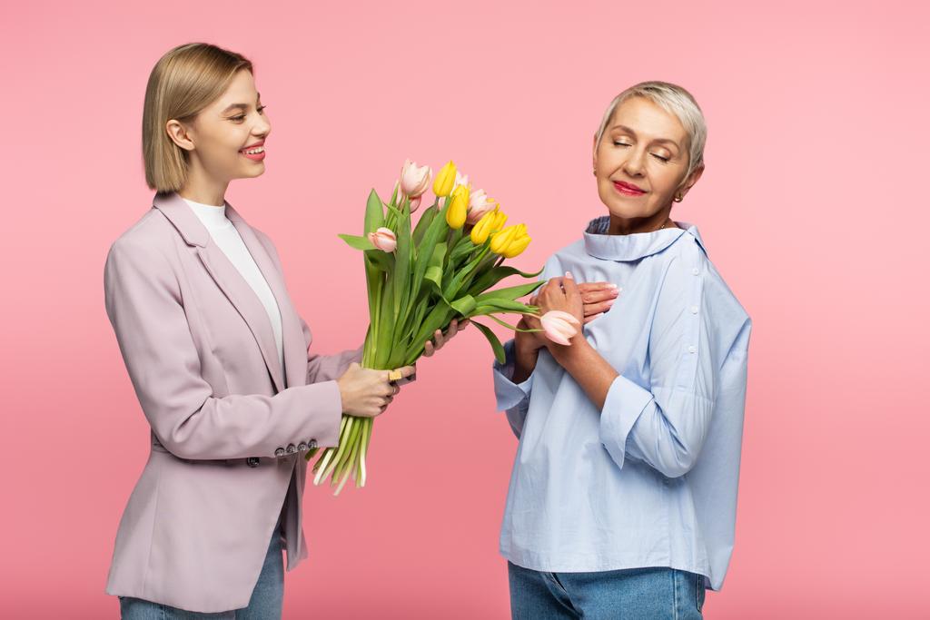 glückliche junge Tochter gibt Blumenstrauß an reife Mutter isoliert auf rosa - Foto, Bild