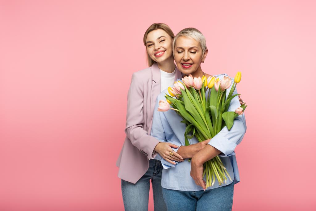 pleased daughter hugging happy middle aged mother holding bouquet of flowers isolated on pink  - Photo, Image