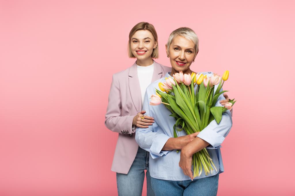 cheerful daughter hugging happy middle aged mother holding bouquet of flowers isolated on pink  - Photo, Image