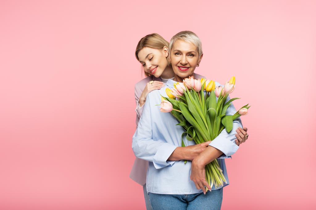 alegre hija abrazando feliz de mediana edad madre sosteniendo tulipanes aislados en rosa - Foto, Imagen