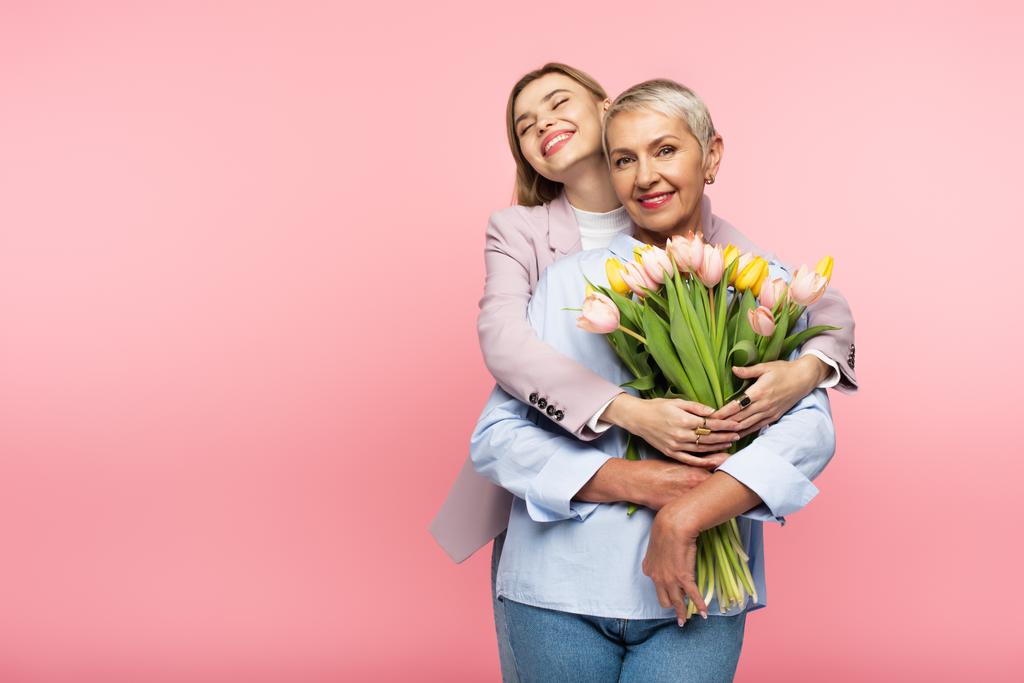 figlia positiva abbracciando felice madre di mezza età che tiene i tulipani isolati su rosa - Foto, immagini