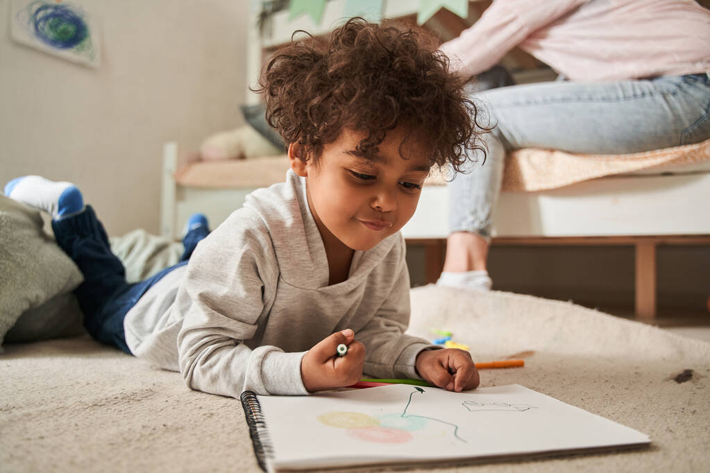 Маленький многорасовый мальчик лежал на полу и рисовал что-то карандашами на бумаге - Фото, изображение