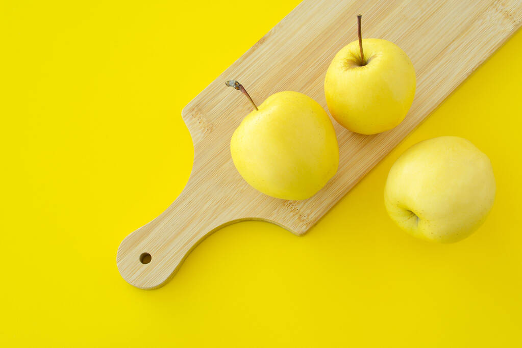 Kolme keltaista omenaa puisella leikkuulaudalla keltaisella pohjalla. Tuoreet kypsät mehukkaat keltaiset hedelmät - Valokuva, kuva