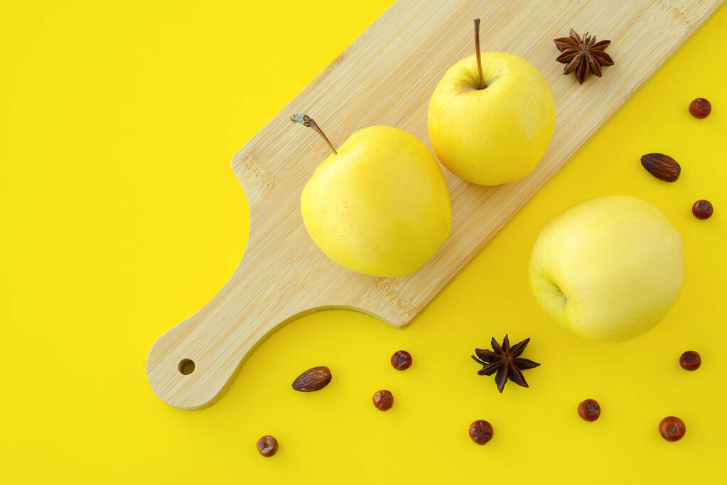 Gelbe Äpfel auf einem Holzschneidebrett vor gelbem Hintergrund, Nüsse, Wacholder, Beeren, Anissterne - Foto, Bild