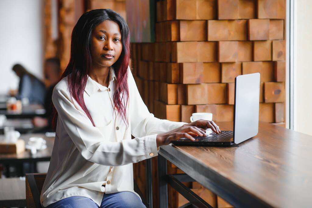 Африканская бизнес-леди ждет встречи в коворкинге и сидит с портативным компьютером. Девушка-хипстер бродит по интернету на ноутбуке во время обеда. - Фото, изображение
