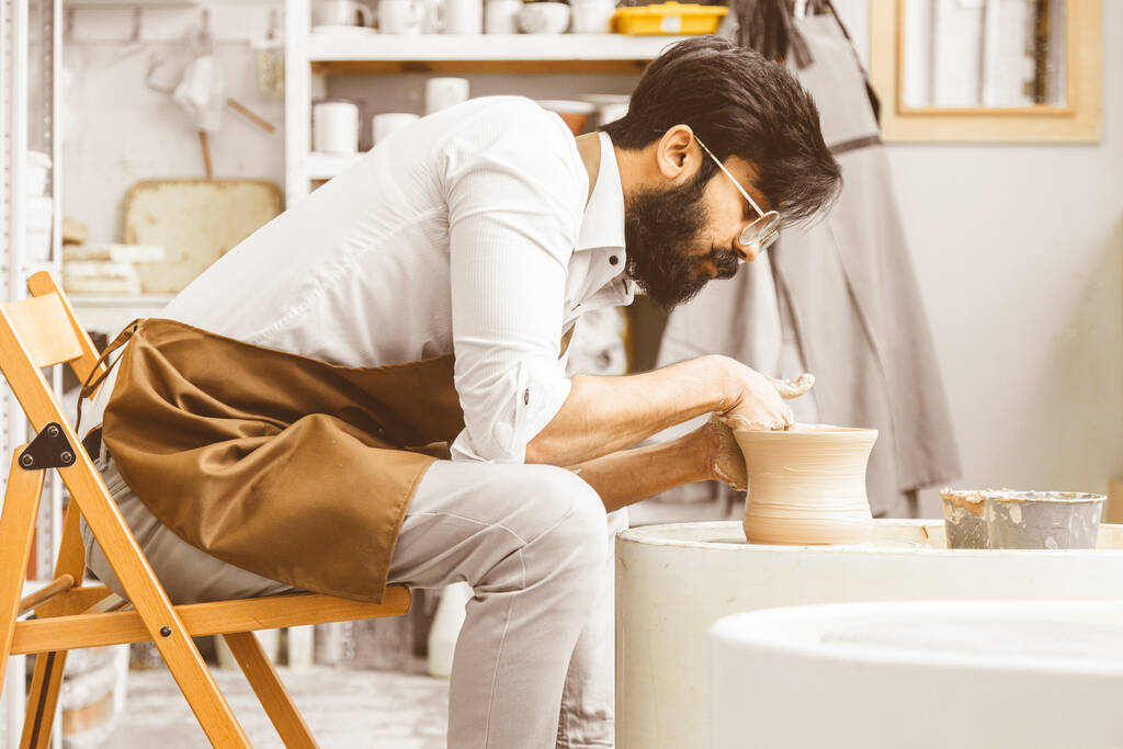 Un alfarero joven se dedica a la artesanía en su taller en una rueda de alfareros y hace un producto de arcilla - Foto, imagen