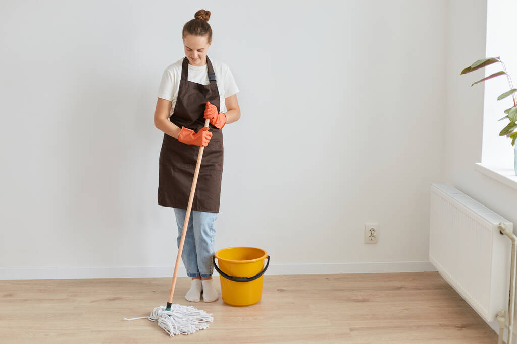 Ganzkörperporträt einer Frau mit orangefarbenen Gummihandschuhen, brauner Schürze und Jeans, die zu Hause den Boden wäscht, lächelnd nach unten schaut und am Wochenende Hausarbeiten erledigt. - Foto, Bild