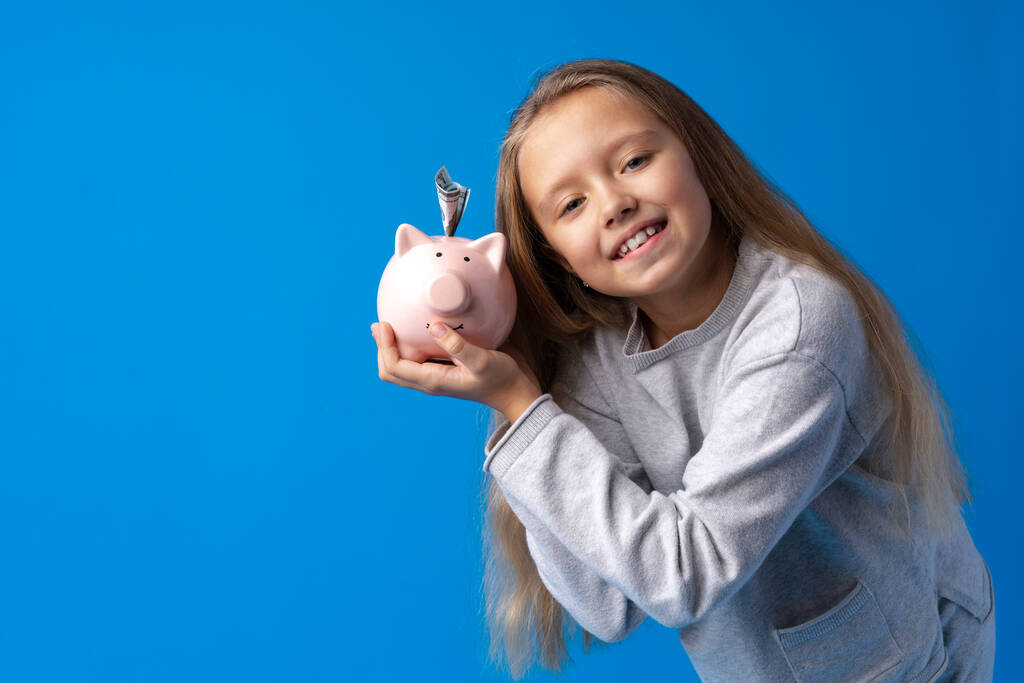schönes kleines Mädchen mit Sparschwein auf blauem Hintergrund - Foto, Bild