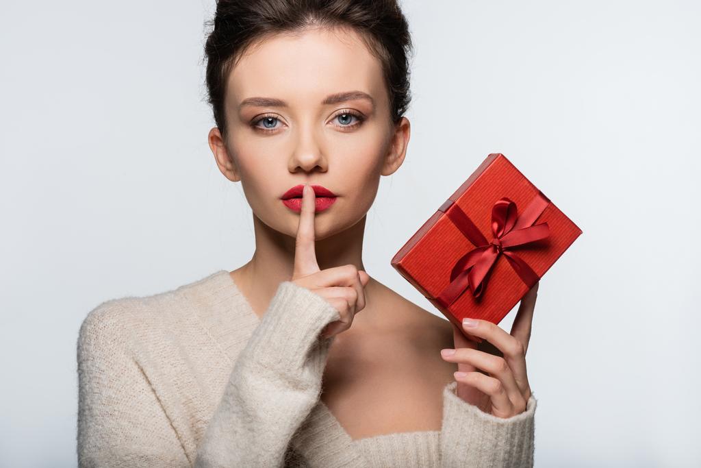 Woman in sweater holding red gift and showing secret gesture isolated on white  - Photo, Image