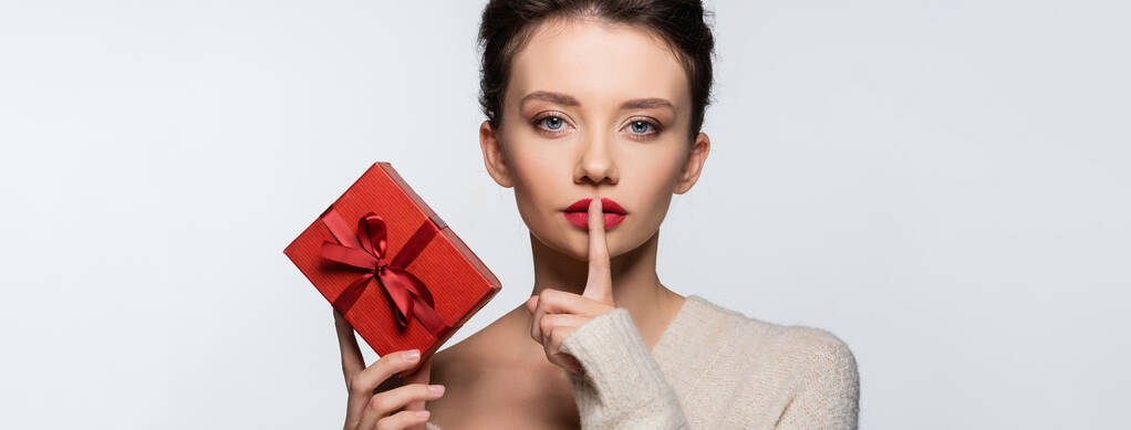 Μελαχρινή γυναίκα δείχνει μυστική χειρονομία και κρατώντας κόκκινο κουτί δώρου απομονωμένο σε λευκό, banner  - Φωτογραφία, εικόνα