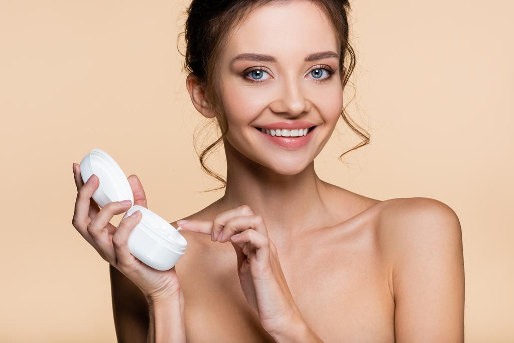 Femme souriante aux épaules nues tenant la crème cosmétique et regardant la caméra isolée sur beige  - Photo, image