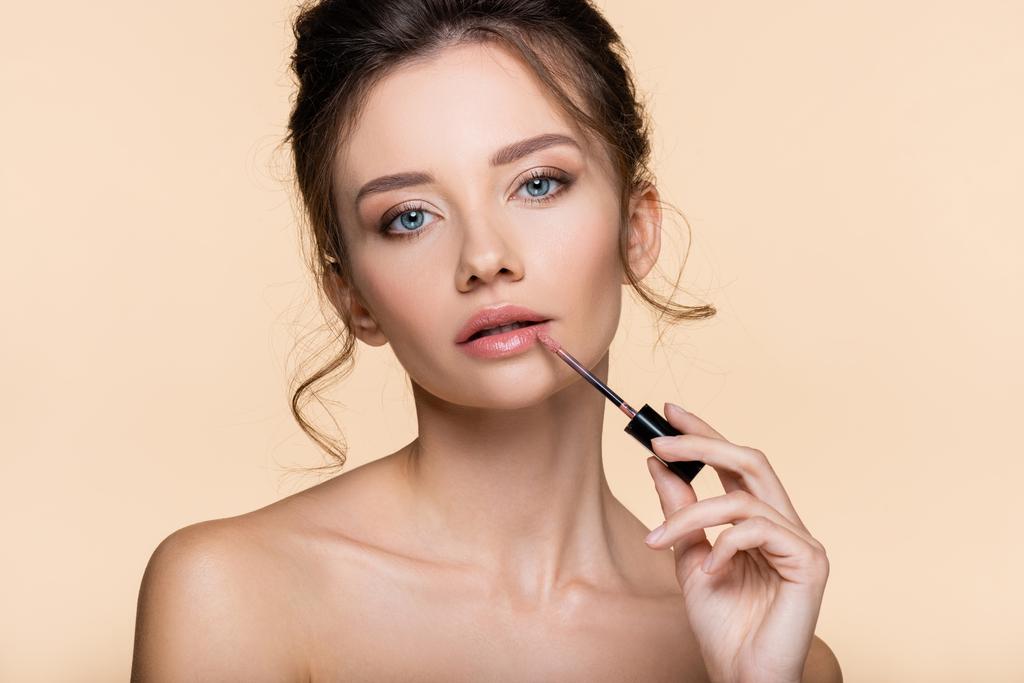 Νεαρή γυναίκα με γυμνούς ώμους εφαρμογή lip gloss απομονώνονται σε μπεζ  - Φωτογραφία, εικόνα