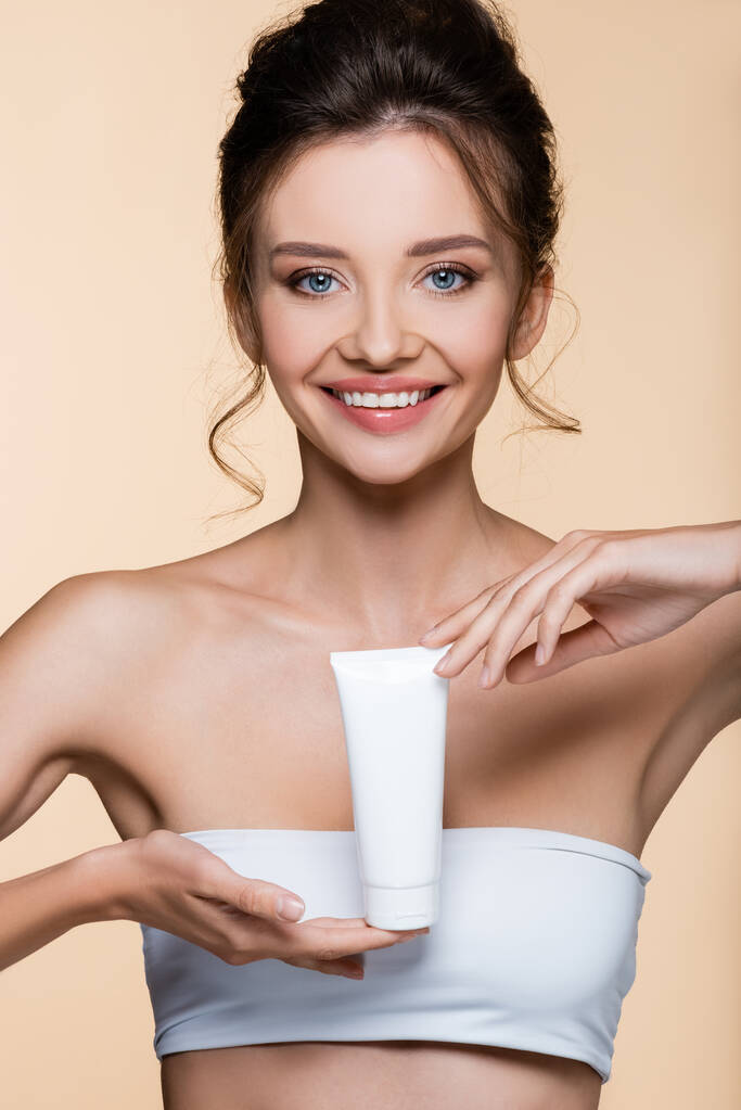 Smiling brunette woman in white top holding cosmetic lotion isolated on beige  - Photo, Image