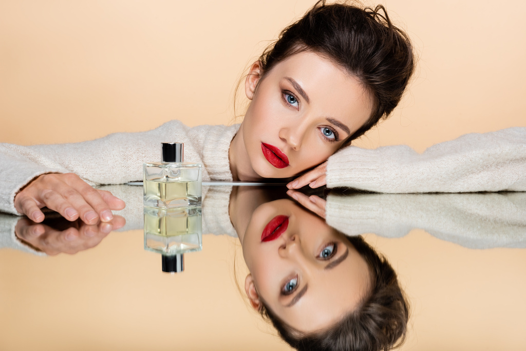 Brunette model met rode lippen kijkend naar camera nabij parfum op spiegel geïsoleerd op beige  - Foto, afbeelding