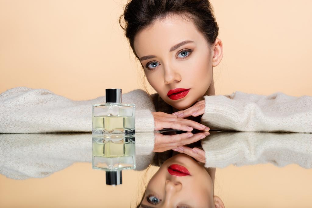 Jovem elegante em camisola quente posando perto de frasco de perfume no espelho isolado no bege  - Foto, Imagem