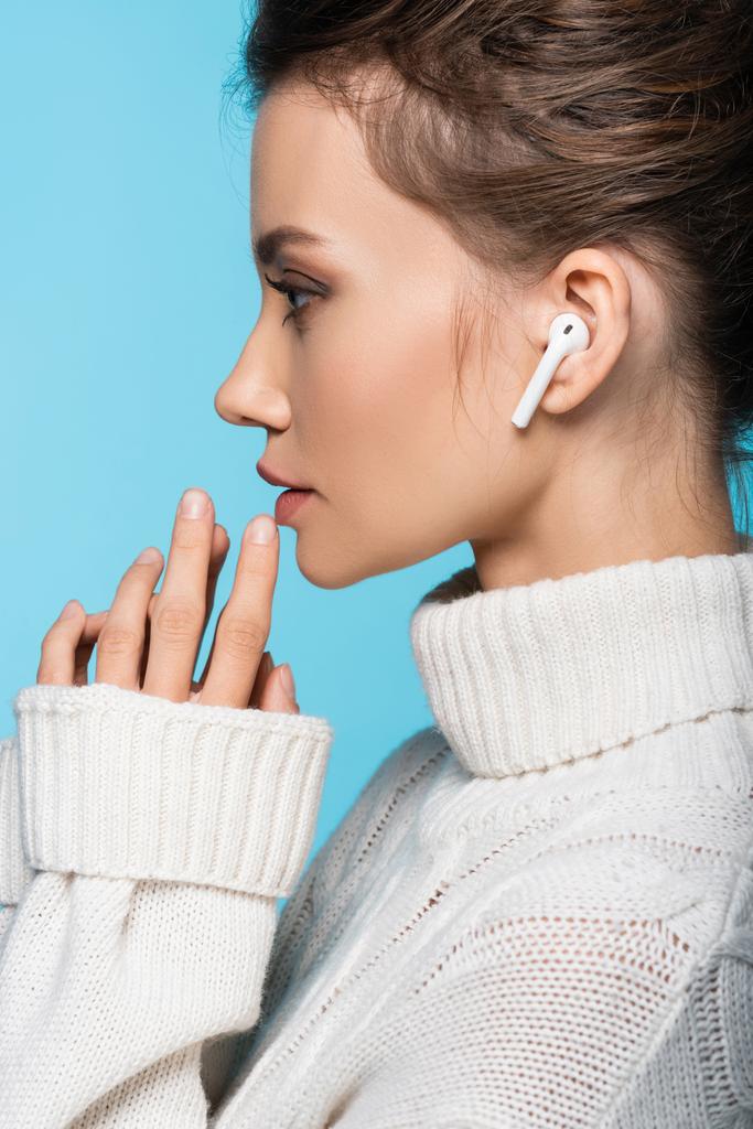 Side view of brunette woman in wireless earphone and sweater isolated on blue - Photo, Image