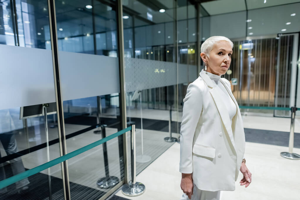 Beyaz takım elbiseli yaşlı bayan modern otelin girişinde duruyor. - Fotoğraf, Görsel