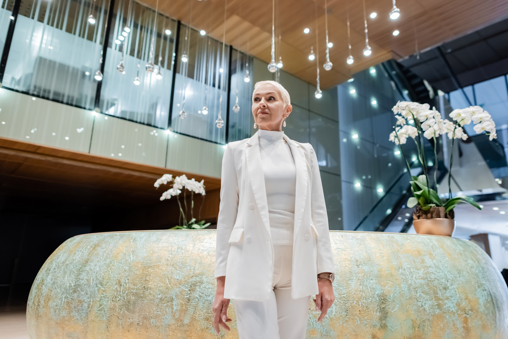 loira empresária sênior em roupas brancas em pé perto de orquídeas em hotel de luxo - Foto, Imagem