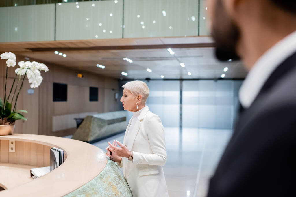 trendy senior businesswoman near hotel reception and bi-racial bodyguard on sturred foreground - Fotografie, Obrázek