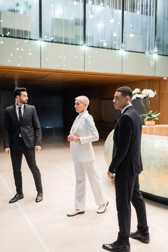 vue pleine longueur de femme d'affaires mature et à la mode marchant près de gardes du corps interracial privés dans le hall de l'hôtel - Photo, image
