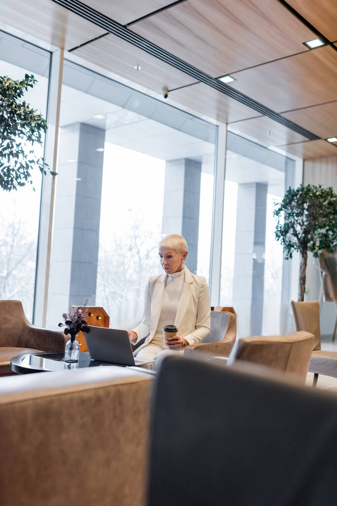senior business lady with takeaway drink using laptop in hotel lounge - Photo, Image