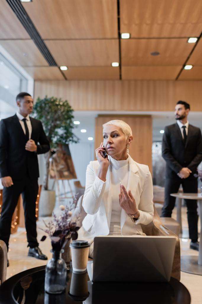 senior businesswoman talking on smartphone near laptop and blurred multiethnic bodyguards - Photo, Image