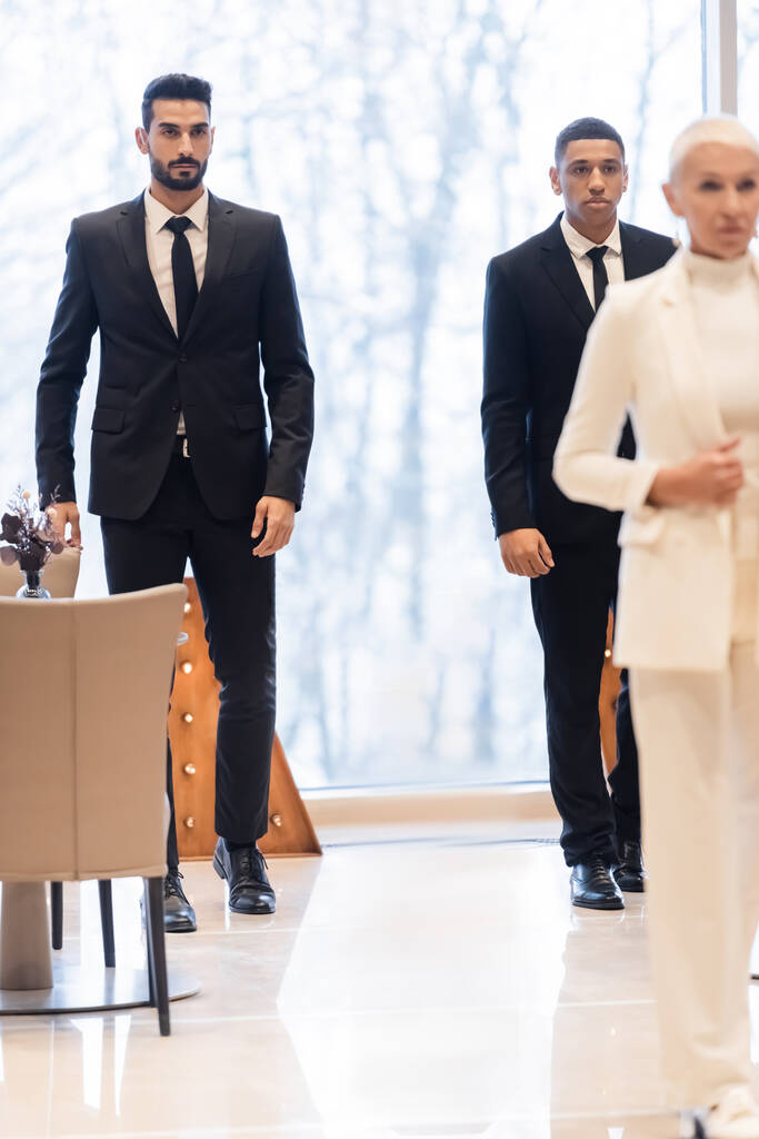 πολυεθνικοί σωματοφύλακες με κοστούμια που περπατούν στο φουαγιέ του ξενοδοχείου κοντά σε ανώτερες επιχειρηματίες σε θολή πρώτο πλάνο - Φωτογραφία, εικόνα