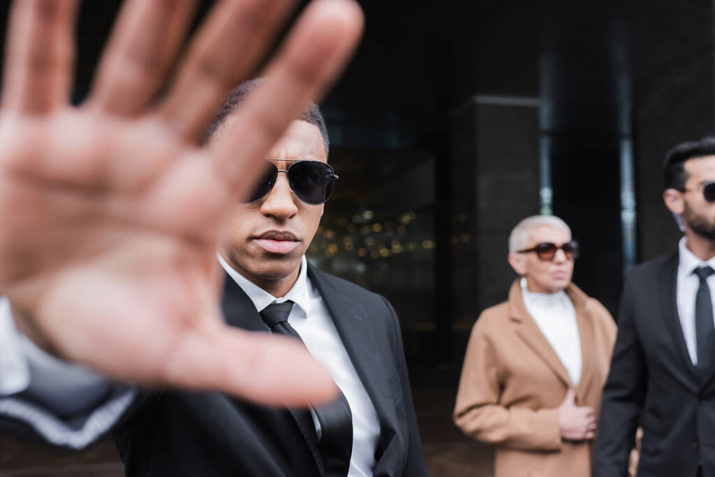 hombre de seguridad de raza mixta mostrando gesto de stop cerca de la mujer de negocios senior y guardaespaldas afroamericano  - Foto, Imagen