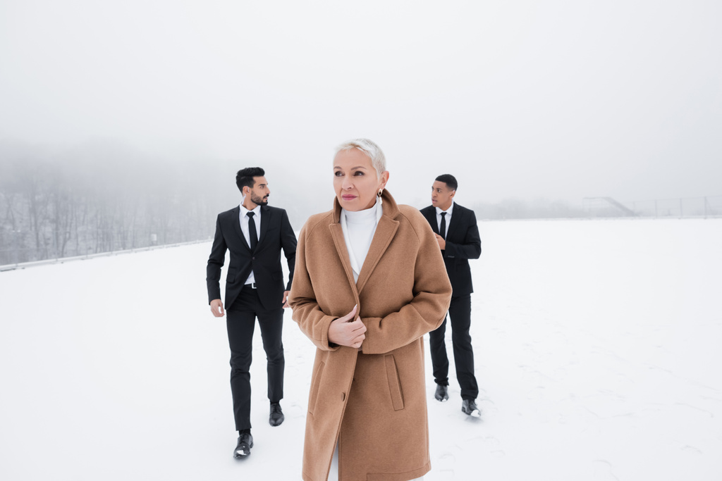 femme d'affaires senior en manteau marchant à travers le champ d'hiver près de gardes du corps multiethniques en costumes - Photo, image