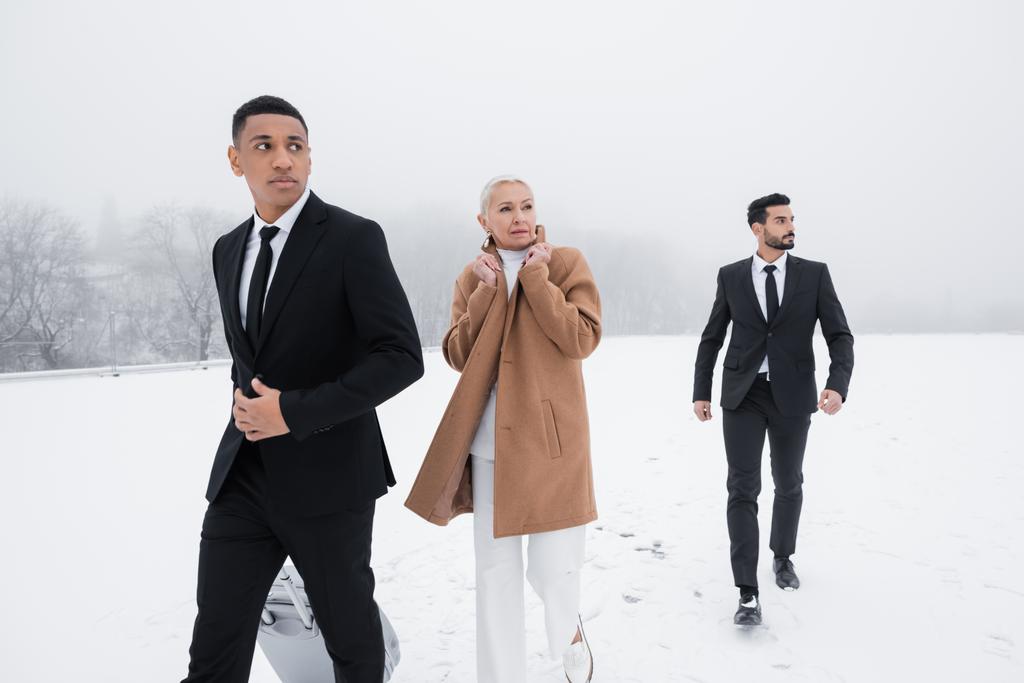 multicultural guards in black suits protecting senior business lady while walking on winter field - Photo, Image