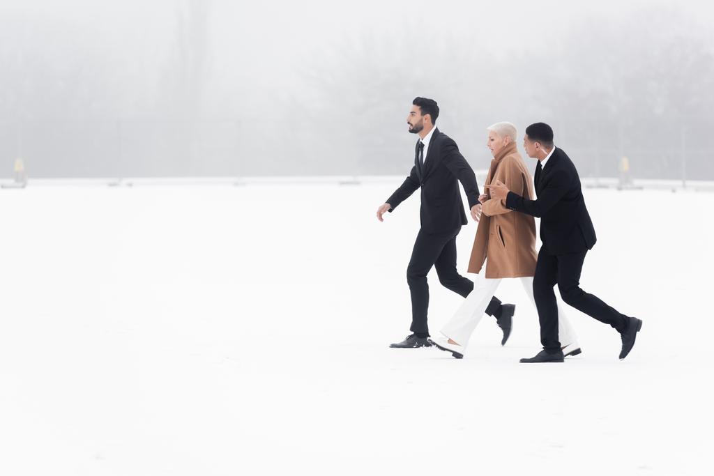 boční pohled na senior businesswoman chůze přes zasněžené pole pod ochranou mezirasových bodyguardů - Fotografie, Obrázek