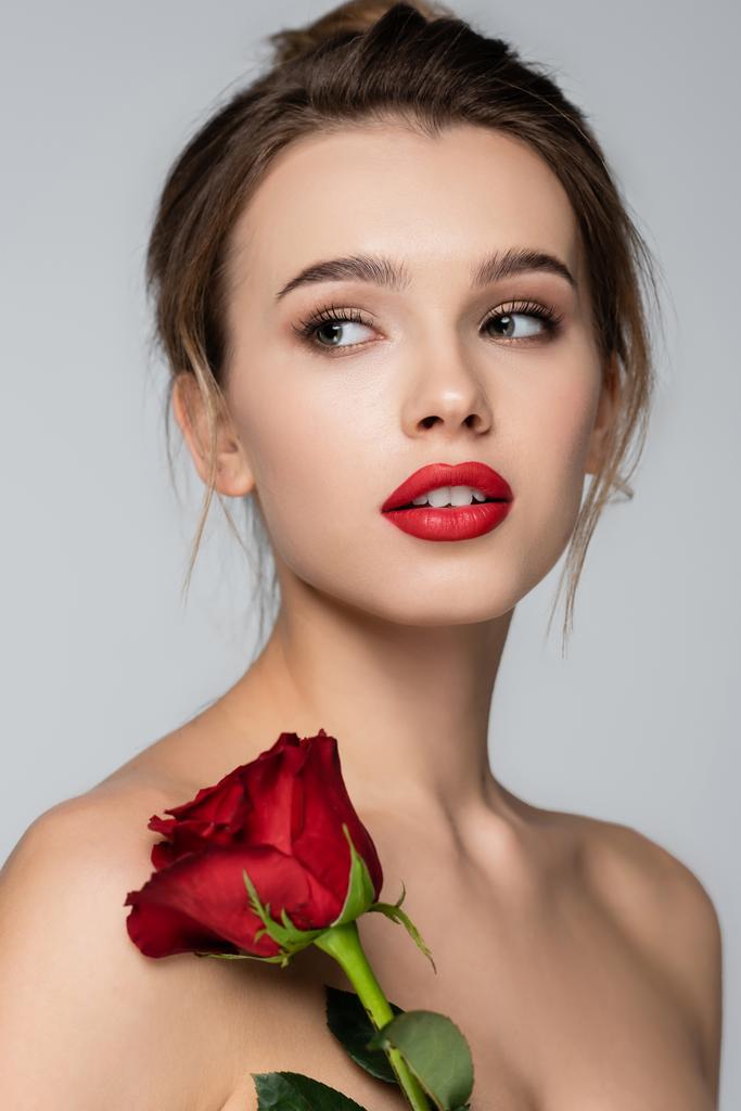 νεαρή γυναίκα με κόκκινα χείλη και τέλειο δέρμα κοιτάζοντας μακριά κοντά στο κόκκινο τριαντάφυλλο που απομονώνονται σε γκρι - Φωτογραφία, εικόνα