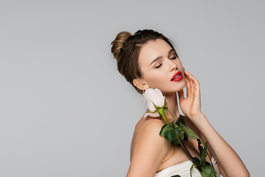 όμορφη γυναίκα με κλειστά μάτια αγγίζοντας το πρόσωπο, ενώ κρατώντας λευκό τριαντάφυλλο απομονωμένο σε γκρι - Φωτογραφία, εικόνα