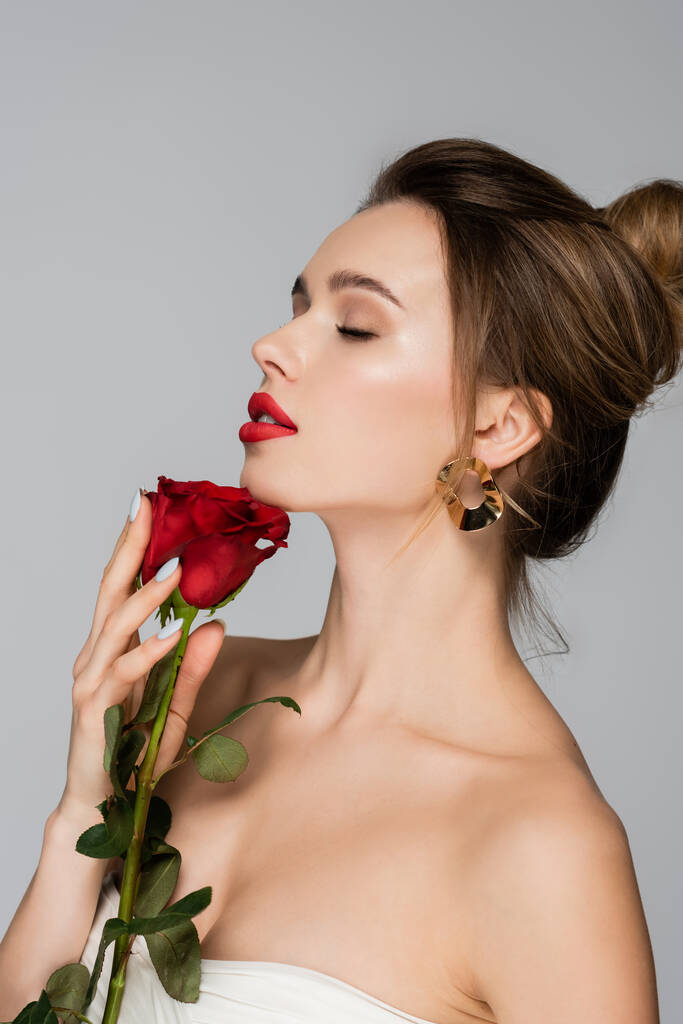 czarująca kobieta z nagimi ramionami i czerwonymi ustami trzymająca różę odizolowaną na szaro - Zdjęcie, obraz