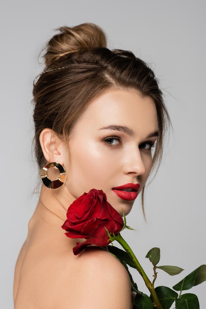 csinos fiatal nő piros ajkak és rózsa közelében csupasz váll nézi kamera elszigetelt szürke - Fotó, kép