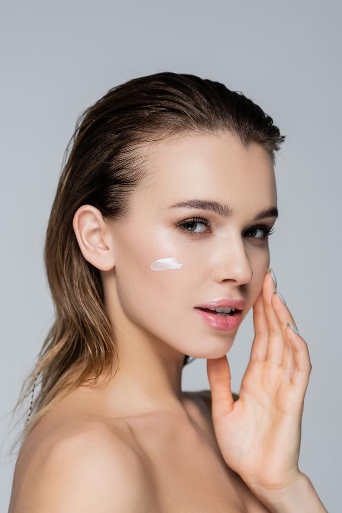jonge vrouw met cosmetische crème op perfect gezicht op zoek naar camera geïsoleerd op grijs - Foto, afbeelding