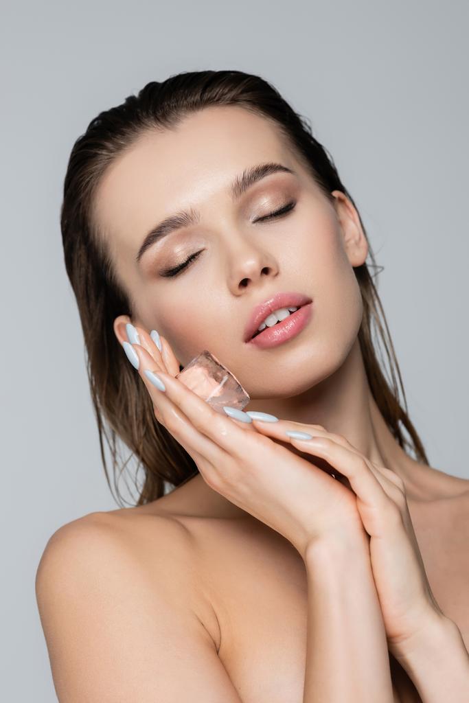 young woman with closed eyes holding ice cube near clean face isolated on grey - Photo, Image