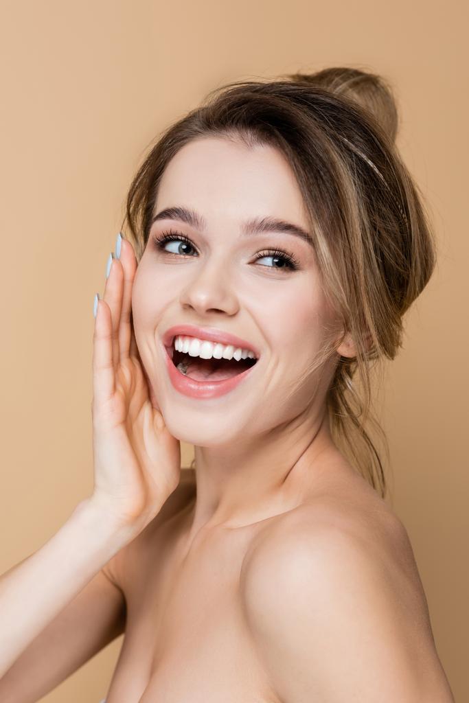 jonge vrouw met natuurlijke make-up aanraken gezicht en lachen geïsoleerd op beige - Foto, afbeelding