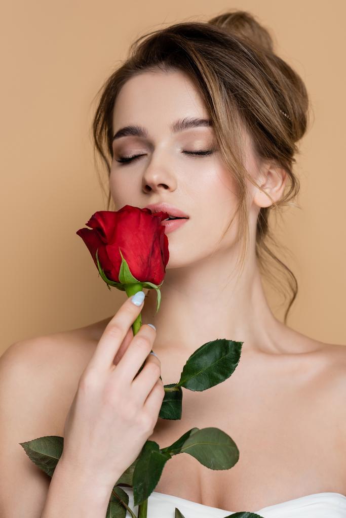 urocza kobieta z gołymi ramionami trzymając czerwoną różę blisko warg odizolowanych na beżu - Zdjęcie, obraz