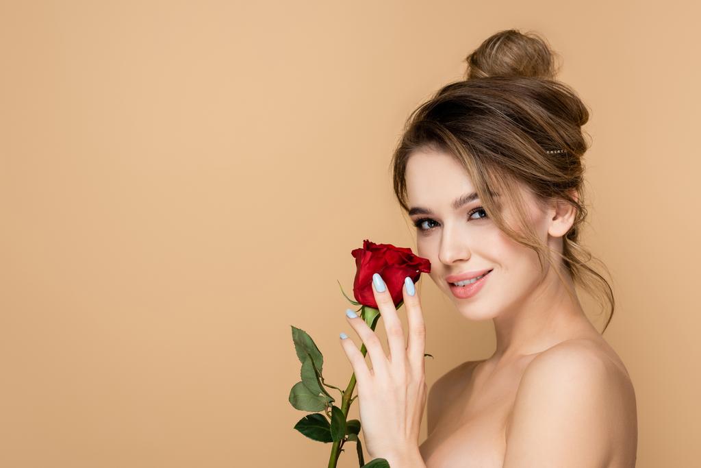 ευχαριστημένη γυναίκα με φυσικό μακιγιάζ και κόκκινο τριαντάφυλλο κοιτάζοντας κάμερα απομονωμένη σε μπεζ - Φωτογραφία, εικόνα
