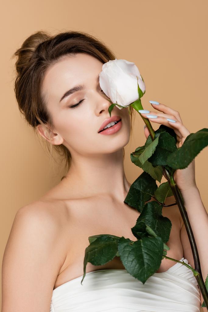 mladá žena s přírodní make-up drží bílou růži v blízkosti obličeje izolované na béžové - Fotografie, Obrázek