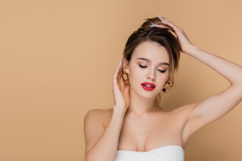 pretty woman in strapless top and golden earrings fixing hair isolated on beige - Photo, Image
