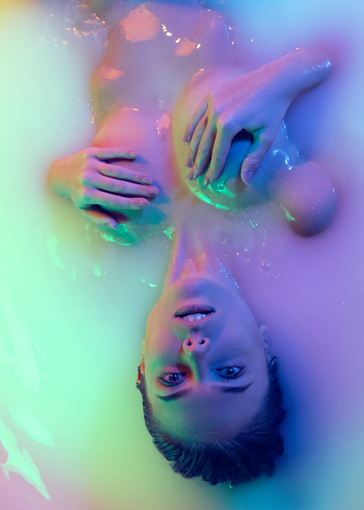 Top view of attractive young girl in milk bath with soft glowing in blue-pink neon light. Copyspace for advertising. Beauty, fashion, style, skincare concept - Photo, Image