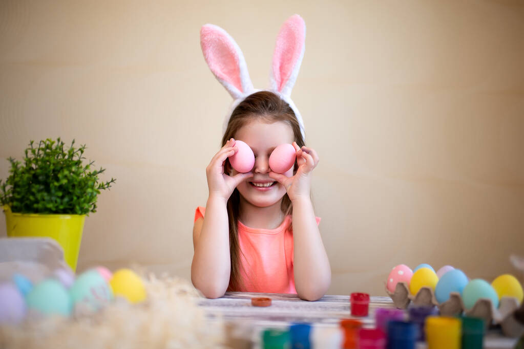 Забавный ребенок держит пасхальные яйца рядом с глазами, готовясь к Пасхе носить кроличьи уши.  - Фото, изображение