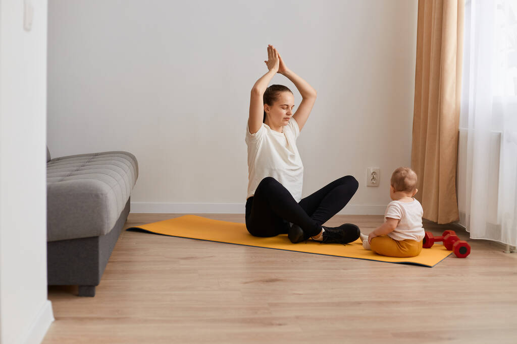 Plan intérieur de la jeune mère fait des exercices de yoga physique avec son bébé tout en étant assis sur un tapis avec les bras levés avec les paumes ensemble, se détendre pendant la dure journée de routine. - Photo, image