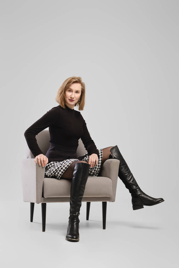 Surprised woman in turtleneck and suspender belt over little skirt sitting in armchair in studio over grey background - Photo, Image