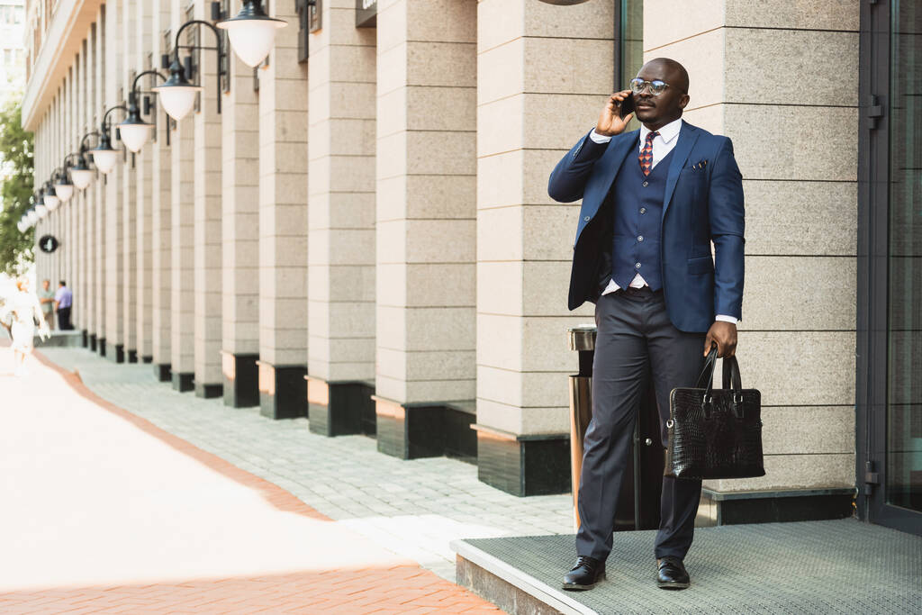 portret czarnego afrykańskiego biznesmena w garniturze na świeżym powietrzu z walizką w ręku stojącą i rozmawiającą przez telefon - Zdjęcie, obraz