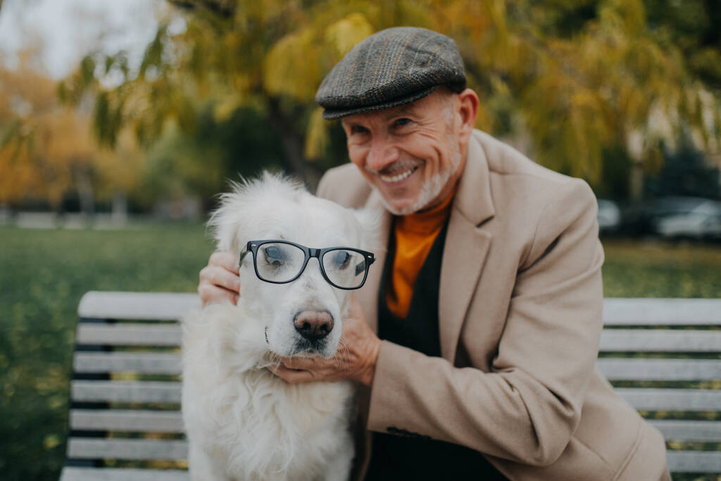 Feliz hombre mayor mirando a la cámara y abrazando a su perro con gafas en el banco al aire libre en la ciudad. - Foto, imagen